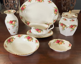 May include: A collection of white porcelain tableware with a floral rose pattern and gold trim. The set includes a large oval platter, a gravy boat, a sugar bowl, a vase, and two bowls. The roses are red, pink, and yellow.