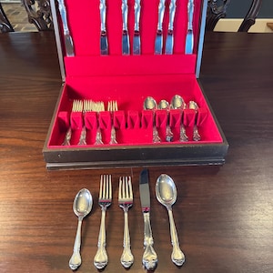 Könnte beinhalten: Eine hölzerne Besteckkiste mit rotem Samtinterieur, die verschiedene silberne Utensilien enthält. Das Set umfasst Messer, Gabeln, Löffel und Servierteile. Einige Teile werden außerhalb der Kiste auf einer Holzoberfläche ausgestellt.