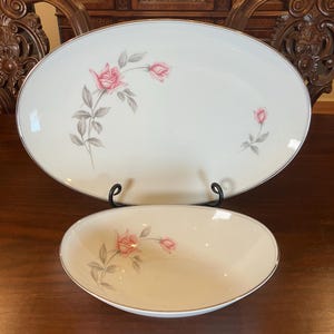 以下が含まれることがあります： 楕円形の皿と小さな楕円形のボウルの2点セット。どちらもオフホワイトで、シルバーのトリムとピンクのバラの花柄が施されています。セットは濃い木製の表面に表示されています。