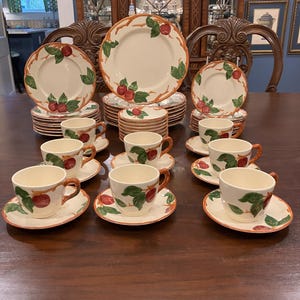 Puede incluir: Un juego de vajilla completo con platos, platillos y tazas de color crema. Cada pieza está adornada con un diseño de manzanas rojas y hojas verdes sobre una rama marrón. El juego incluye varios tamaños de platos y tazas de té.