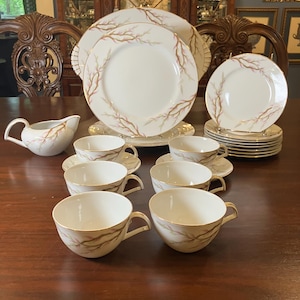 Op de afbeelding: Een complete porseleinen eetservies met witte borden, kopjes, schotels en een melkkan. Elk stuk heeft een bruin en roze takkenpatroon met gouden randen. De set wordt gepresenteerd op een donkere houten tafel.
