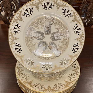 May include: A stack of cream-colored decorative plates with gold trim and intricate designs. The plates feature a central image of figures and a border with cut-out patterns. The plates are stacked on a dark wooden surface.