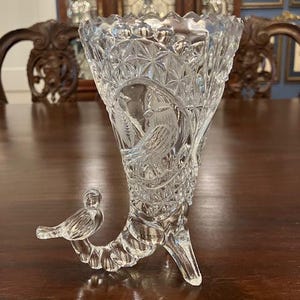 May include: Clear glass vase with a horn shape and a bird-shaped handle. The vase features intricate cut glass patterns, including stars and bird motifs. The vase has three feet and a scalloped rim, showcasing a vintage aesthetic.