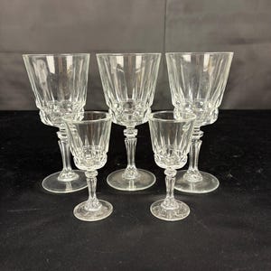 Op de afbeelding: Een set van vijf heldere glazen op voet, bestaande uit drie grotere glazen en twee kleinere glazen. Elk glas heeft een gefacetteerde kelk met verticale ribbels, een korte steel en een ronde voet, allemaal gemaakt van transparant glas.