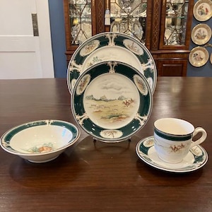 Op de afbeelding: Een set van vier witte porseleinen serviesstukken met een groene en gouden rand. Het servies heeft een jachtscène met paarden en ruiters. De set bevat een kom, een bord, een schoteltje en een theekopje.