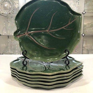 Könnte beinhalten: Ein Set aus sechs grünen, blattförmigen Keramikplatten. Die Teller haben eine glänzende Oberfläche mit braunen Aderdetails. Ein Teller wird auf einem schwarzen Metallständer präsentiert. Die Teller sind gestapelt.