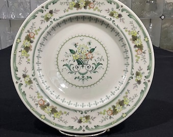 Puede incluir: Un plato de cena de cerámica blanca con un diseño floral verde y amarillo. El plato tiene un borde elevado y un diseño central de una cesta de flores.
