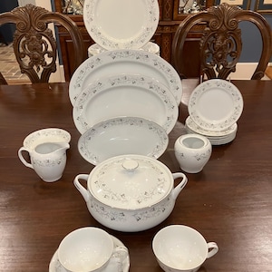 Puede incluir: Un juego completo de vajilla de porcelana blanca con un delicado estampado floral gris y ribete plateado. El juego incluye platos, cuencos, tazas, platillos, una fuente para servir, una cacerola cubierta, una lechera y una azucarera.