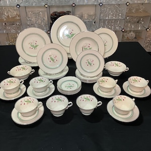 May include: A collection of white porcelain dinnerware with a floral design. The set includes plates, saucers, and teacups with a green and gold trim. The floral pattern features pink and green accents. The arrangement is on a black surface.