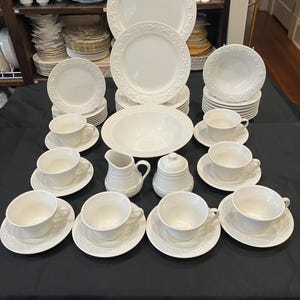 Pode incluir: Um conjunto de louça de porcelana branca, incluindo pratos, tigelas, xícaras, pires, um cremeira e um açucareiro. As peças têm um desenho floral em relevo nas bordas. O conjunto está disposto sobre uma superfície preta.