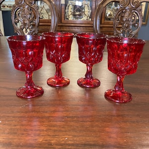 May include: Four ruby red stemmed glasses with a diamond pattern. The glasses are arranged on a dark wooden table. The glasses are approximately 4 inches tall.