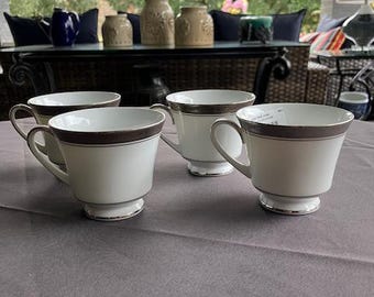 May include: Four white teacups with a silver rim and a silver band around the middle. The teacups are on a gray tablecloth.