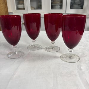 May include: Four ruby red wine glasses with clear stems and bases are arranged on a white tablecloth. The glasses have a classic, elegant shape, and the rich color of the glass is visible. The background includes a white cabinet.