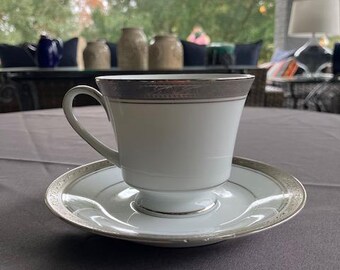 May include: A white teacup and saucer with a silver trim. The cup has a delicate design and the saucer has a scalloped edge.