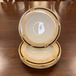 May include: A stack of beige and white ceramic bowls with a black and gold border. The bowls are stacked on a metal stand, showcasing their decorative design. The bowls have a classic, elegant aesthetic.