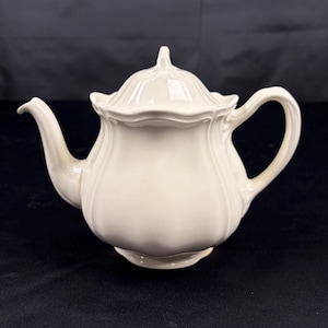 May include: A cream-colored ceramic teapot featuring a scalloped rim, a curved handle, and a matching lid with a pointed finial. The body of the teapot has vertical embossed ridges that extend from the base to the top, creating a fluted appearance.