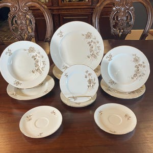 May include: A set of white porcelain dinnerware featuring a floral design in brown and gold. The collection includes plates, bowls, and saucers, displayed on a dark wooden table. The plates have gold trim around the edges.