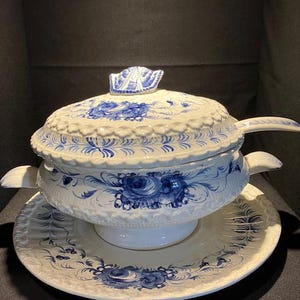 Puede incluir: Un tazón de servir de cerámica blanca con un diseño floral azul y blanco. El tazón tiene una tapa y un plato de servir a juego. El tazón está decorado con un patrón floral en relieve.