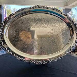 Pode incluir: Uma bandeja de servir oval grande de prata com alças ornamentadas e enroladas. A bandeja tem uma superfície altamente polida e reflexiva com um padrão em relevo complexo.