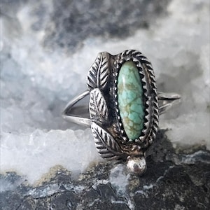 May include: A silver ring featuring an oval-shaped turquoise stone with brown matrix veining. The stone is set in a twisted metal border and accented with three embossed leaf designs on one side and a small silver bead at the base of the setting.