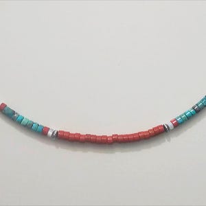 May include: A beaded necklace featuring small, square red beads. Accents of turquoise, white, and silver beads are interspersed throughout the necklace. The necklace is displayed against a plain white background.