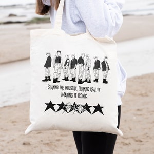 May include: Cream-colored tote bag featuring a black and white illustration of a group of people. Below the illustration are the words "Shaking the Industry, Quaking Reality, Making it Iconic" and five black stars. The bag is held by a person.