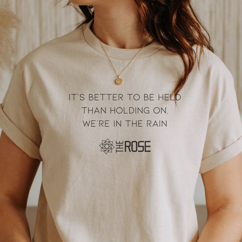 May include: A beige t-shirt with the text "It's better to be held than holding on. We're in the rain" and a logo that says "The Rose" with a geometric design.