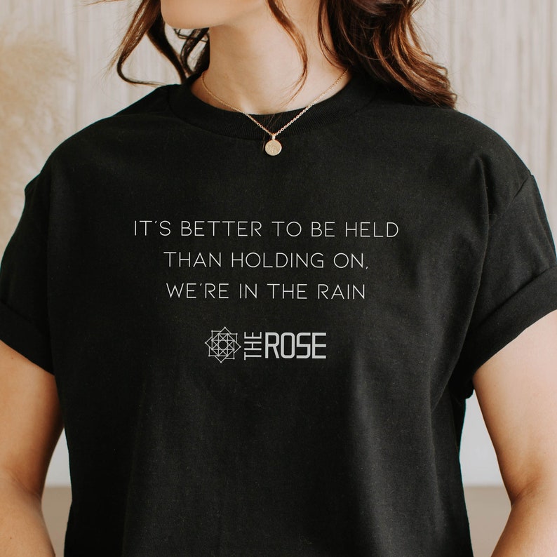May include: Black t-shirt with white text that reads "It's better to be held than holding on, we're in the rain." Below the text is a geometric design with the word "The Rose" inside.