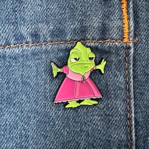 May include: A green cartoon lizard pin wearing a pink dress and a pink collar. The pin is set against a blue denim background, with the lizard's arms outstretched. The pin is a fun accessory.