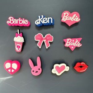 Peut inclure: Un ensemble de dix breloques en plastique rose et bleu avec différents motifs, notamment des cœurs, des nœuds, un lapin, un signe de paix, des lèvres et les mots "Barbie" et "Ken".