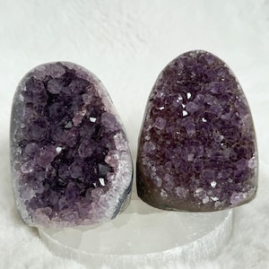 Puede incluir: Dos geodas de amatista, cada una con un interior cristalino de color púrpura oscuro. Las geodas se exhiben sobre una base blanca semitranslúcida, sobre un fondo blanco suave. Las geodas tienen una forma natural y orgánica.