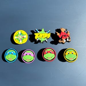 May include: Set of eight Teenage Mutant Ninja Turtles themed shoe charms. The charms feature the four turtles, a pizza, the word "TURTLES" with a turtle shell, and a character from the show.