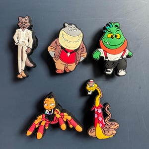 May include: Five cartoon character shoe charms. A wolf wearing a white suit, a hippo wearing a red shirt and brown jacket, a green monster wearing overalls, a spider wearing a red shirt, and a snake wearing a red shirt with a floral pattern.