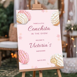 May include: A pink baby shower sign on an easel. The sign features watercolor illustrations of concha pastries and text that reads "There's a little Conchita in the oven!" and "Welcome to Victoria's Babyshower, April 25, 2026."
