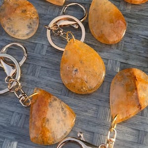 May include: A collection of keychains featuring polished, teardrop-shaped stones in shades of amber, orange, and gold with dark inclusions. Each stone is attached to a circular metal split ring and a lobster claw clasp in a silver tone.