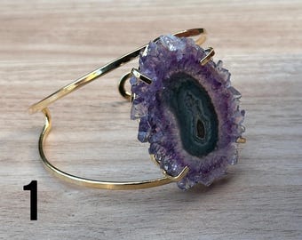 May include: A gold-colored metal cuff bracelet featuring a large, oval-shaped amethyst geode. The geode has a dark center with concentric rings of blue and green, surrounded by a ring of purple crystals. The bracelet is displayed on a light wood surface.