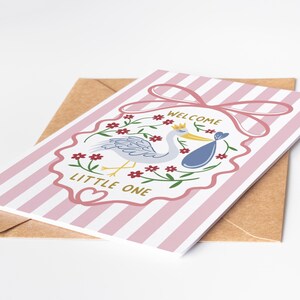May include: A greeting card with pink and white stripes, featuring a stork carrying a baby in a blue bundle. The card has a pink bow and the words "WELCOME LITTLE ONE". It rests on a brown envelope.