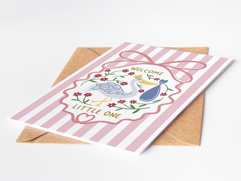 May include: A greeting card with pink and white stripes, featuring a stork carrying a baby in a blue bundle. The card has a pink bow and the words "WELCOME LITTLE ONE". It rests on a brown envelope.