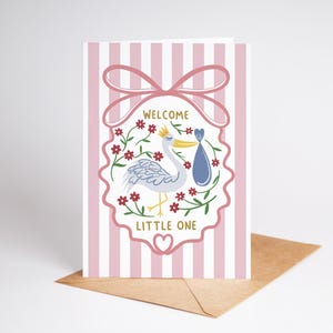 May include: A greeting card with a pink and white striped background. The card features a stork carrying a bundle, surrounded by flowers, and the words "Welcome Little One". A pink bow tops the design, and the card rests on a brown envelope.