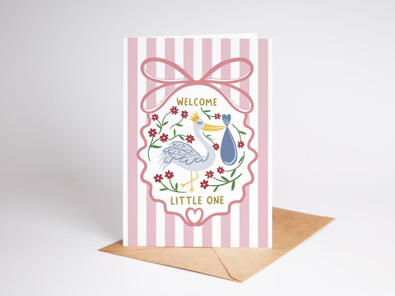 May include: A greeting card with a pink and white striped background. The card features a stork carrying a bundle, surrounded by flowers, and the words "Welcome Little One". A pink bow tops the design, and the card rests on a brown envelope.