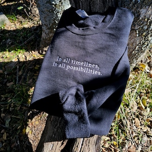Peut inclure: Sweat-shirt noir avec le texte "In all timelines, in all possibilities." brodé en blanc sur le devant.