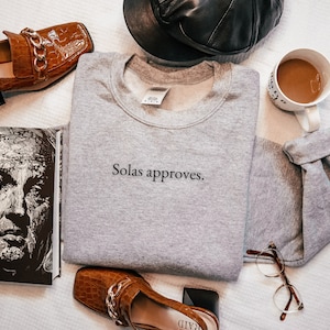 Puede incluir: Una sudadera gris con el texto "Solas approves." impreso en la parte delantera. La sudadera está rodeada de varios artículos, incluyendo un libro, un par de zapatos marrones, una taza de café y un par de gafas.