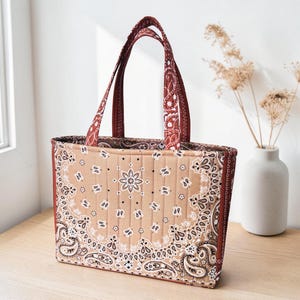 Könnte beinhalten: Eine gesteppte Tragetasche mit einem beigefarbenen und dunkelbraunen Paisley-Bandana-Muster. Die Tasche verfügt über zwei passende Stoffgriffe und eine rechteckige Form mit vertikalen Steppnähten, die sich über das gesamte Stoffmuster ziehen.