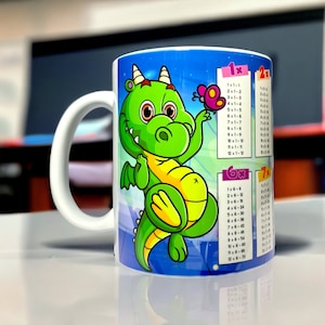 Könnte beinhalten: Eine weiße Keramiktasse mit einer grünen Drachenillustration und einem Schmetterling. Die Tasse zeigt die Multiplikationstabellen von 1x bis 7x.