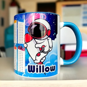 Könnte beinhalten: Weißer Keramikbecher mit blauem Henkel und Rand. Der Becher zeigt ein Cartoon-Astronauten-Design mit dem Namen "Willow" und Multiplikationstabellen. Der Hintergrund hat ein Weltraumthema mit Sternen und Planeten.