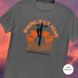 Può includere: T-shirt grigio melange scuro con una grafica arancione di un elettricista che lavora su una linea elettrica con palme e il testo "Making it Lit Again Florida Hurricane Ian 2022".