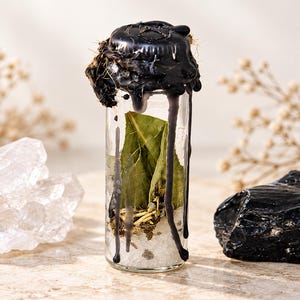 May include: A clear glass vial sealed with black wax that drips down the sides. Inside the vial are green bay leaves, dried herbs, and white salt crystals. The top of the wax seal features a stamped circular emblem with a visible design.