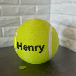 May include: A bright yellow tennis ball-shaped container with the name "Henry" in black lettering. The container has white stripes and a lid. A small coin is next to the container.