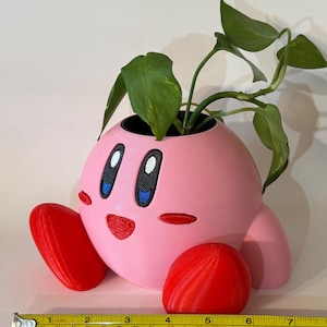 Op de afbeelding: Een roze 3D-geprinte plantenbak in de vorm van een stripfiguur, met rode voeten en een rode bol. De plantenbak bevat een groene bladplant. Een gele rolmaat geeft aan dat de plantenbak ongeveer 15 cm breed is.
