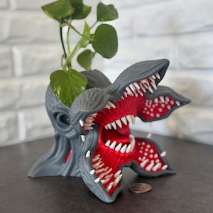 Puede incluir: Una maceta Demogorgon gris con interior rojo y dientes blancos, que contiene una planta verde. La maceta está sobre una superficie oscura con una pared de ladrillos al fondo. Una moneda está al lado de la maceta.
